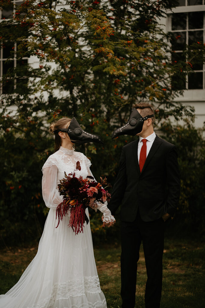 
A couple wearing bird masks holding hands in front of a chapel. 