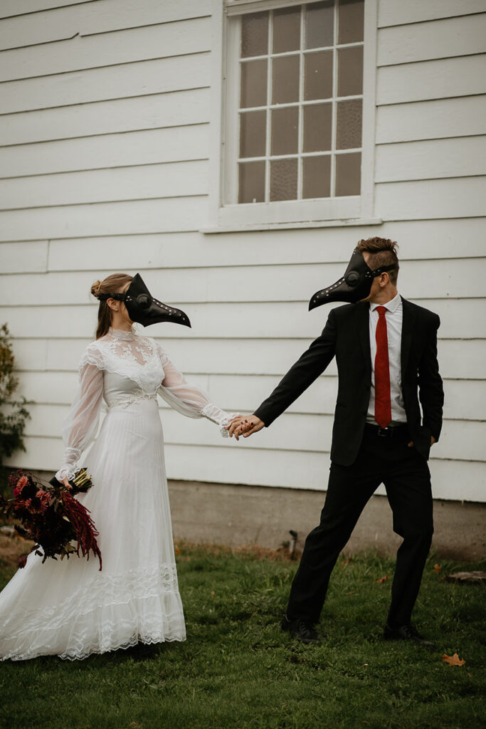A couple wearing bird masks in front of a chapel looking at each other and holding hands. 