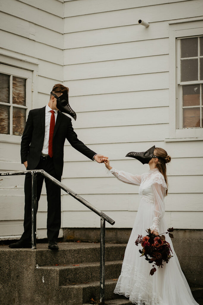 The husband leading his wife up the stairs of a chapel. They both are wearing bird masks. 