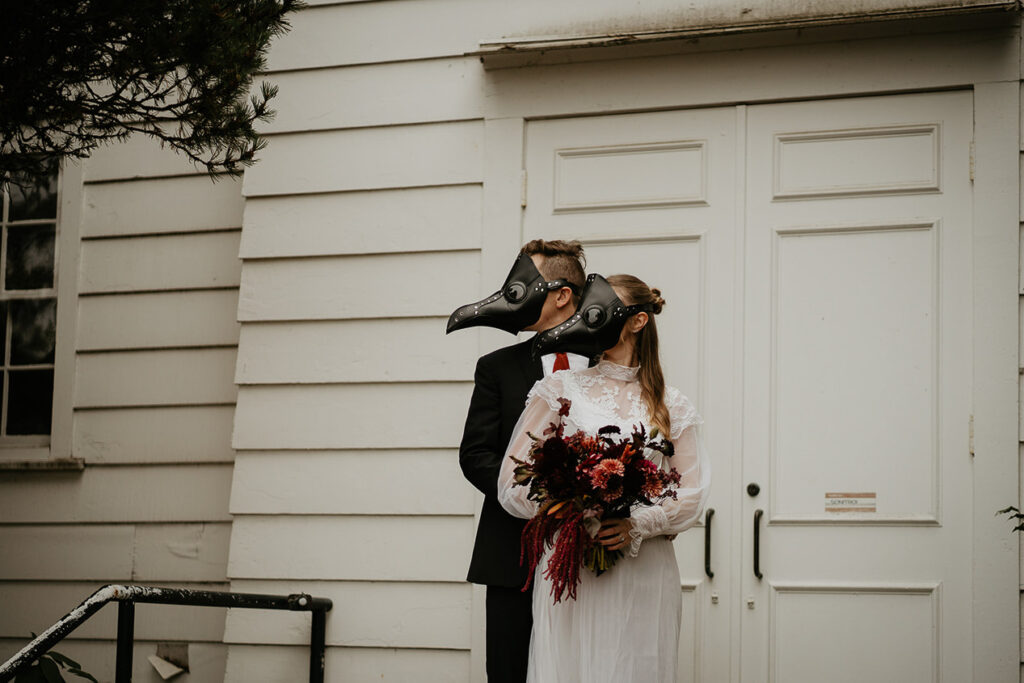 A couple wearing bird masks holding each other in front of a chapel. 