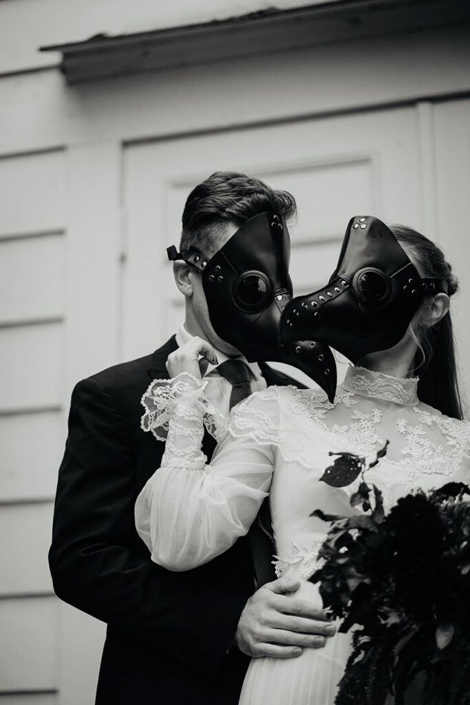 A black and white photo of a couple wearing bird masks holding each other in front of a chapel. 
