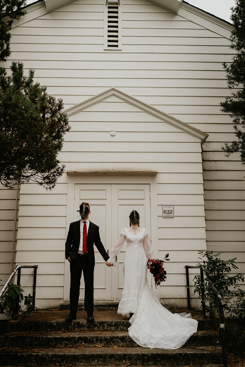 A couple wearing bird masks in front of a chapel holding hands. 