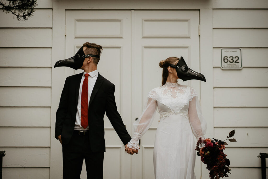 A couple wearing bird masks in front of a chapel holding hands. 