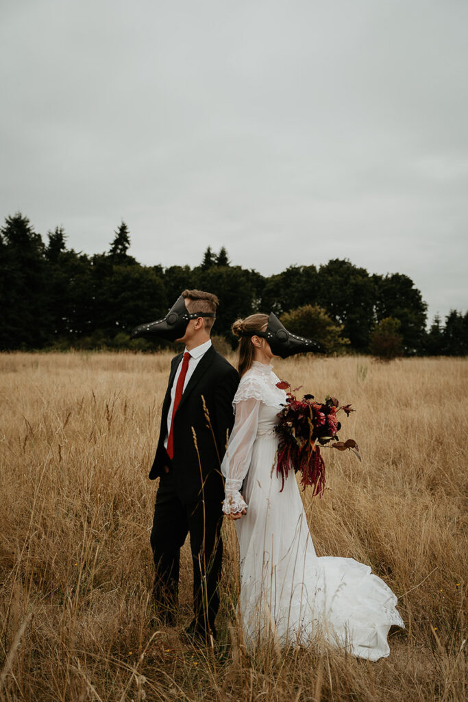 A couple wearing bird masks holding hands standing in an open field. 
