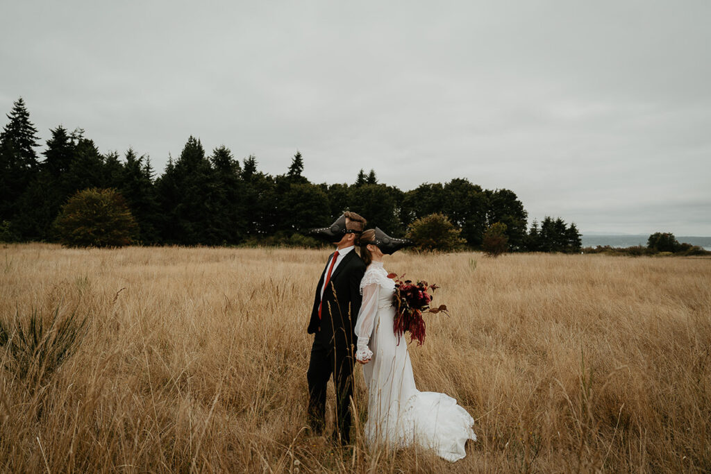 A couple holding hands back to back in a field. They are also wearing bird masks. 
