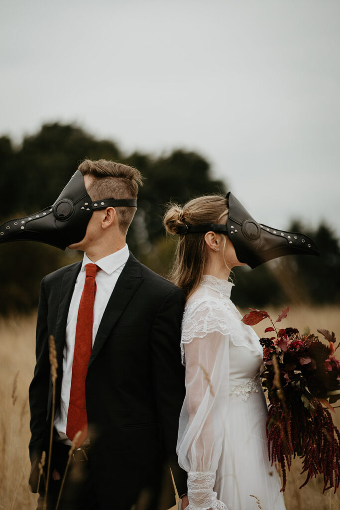 A couple holding hands back to back in a field. They are also wearing bird masks. 