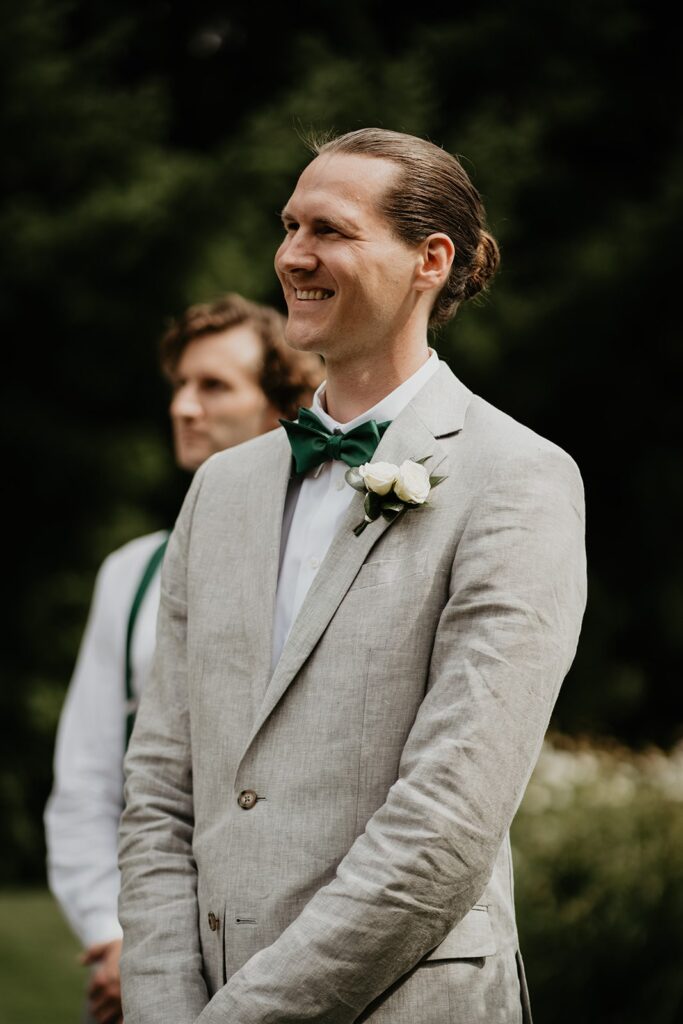 Groom in gray linen suit smiles as he watches bride walk down the wedding aisle