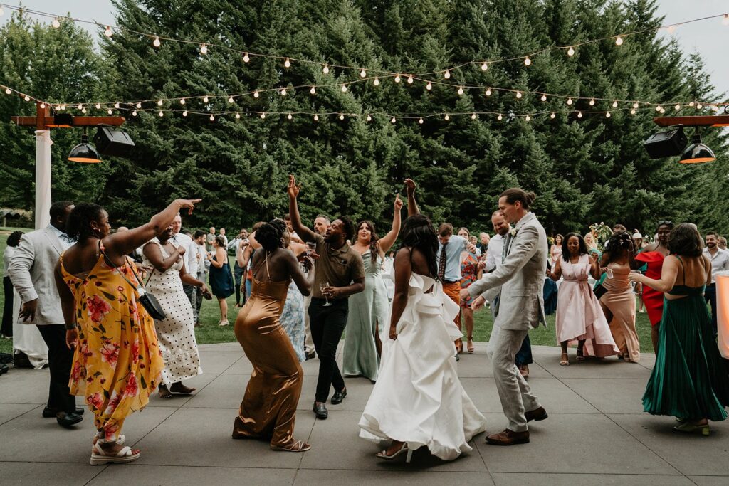 Bride and groom dance with guests under string lights at outdoor wedding reception
