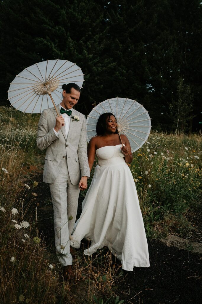 Bride and groom hold parasols while walking through a wildflower field at Gorge Crest Vineyards wedding