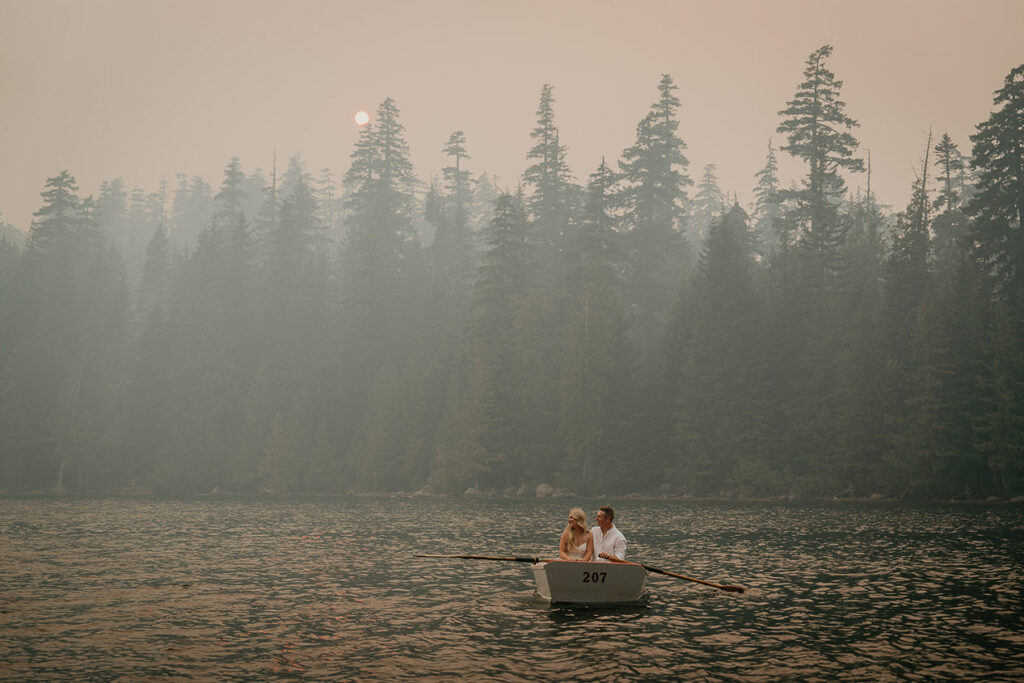 A couple rowing a boat in Lost Lake, with smoke and pine trees in the background. 