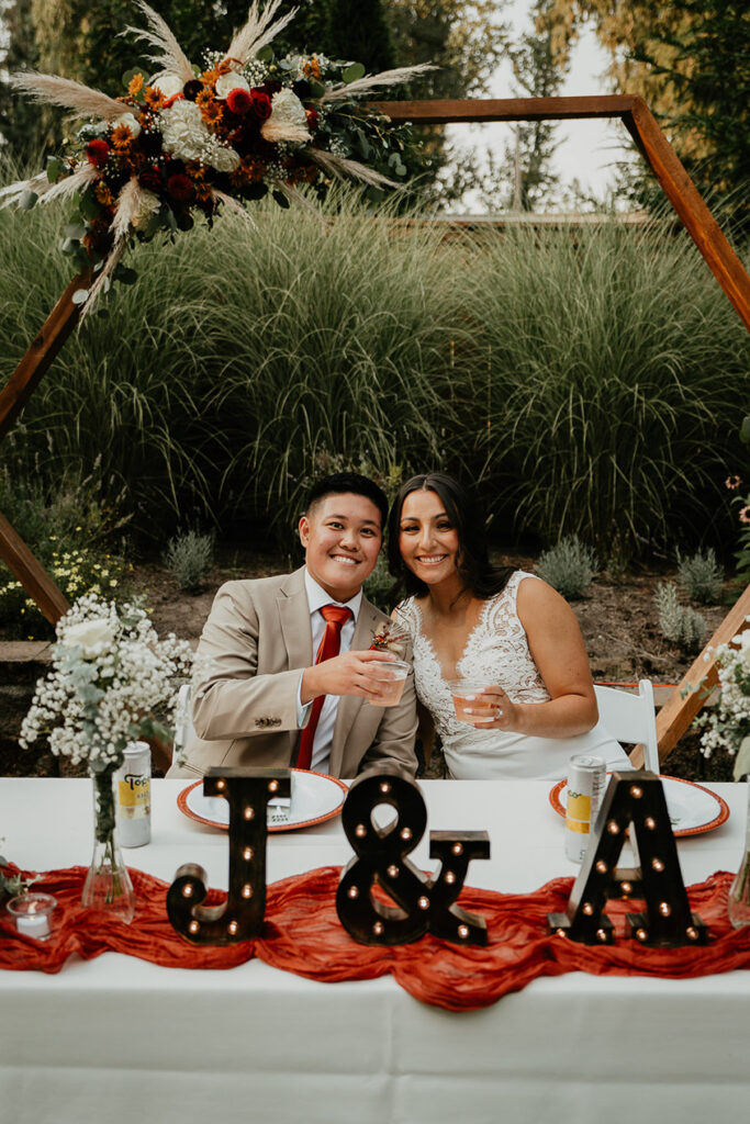 The newlyweds holding drinks and smiling while sitting down at their table. 