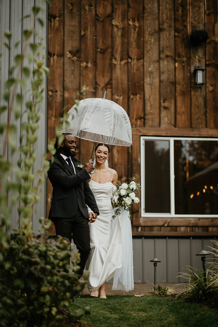 The bride and groom walking to the altar with a clear umbrella to shield them from the rain. 