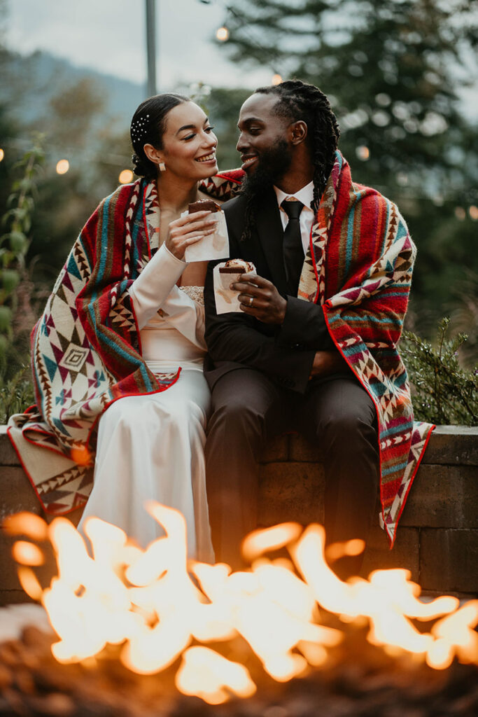 The bride and groom in a blanket, eating s'mores by a fire. 