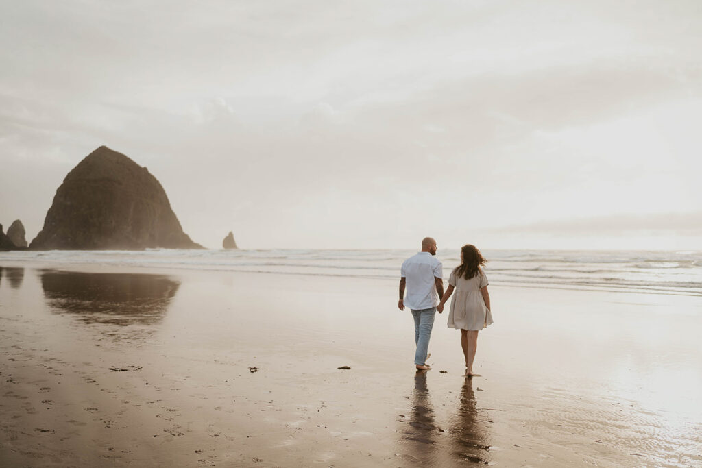 a couple holding hands and walking barefoot in Cannon Beach with Haystack Rock in the background. 