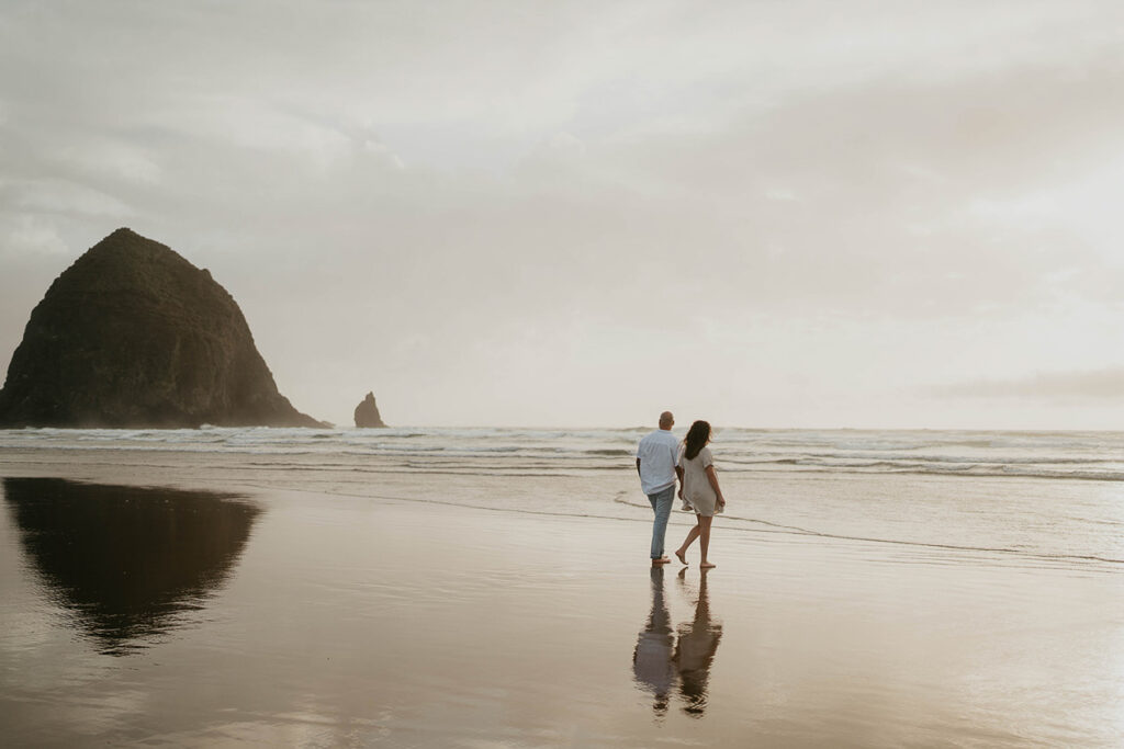 a couple holding hands and walking barefoot in Cannon Beach with Haystack Rock in the background. 