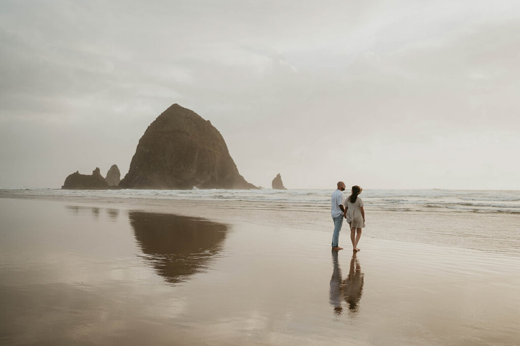 a couple holding hands and walking barefoot with Haystack Rock in the background. 