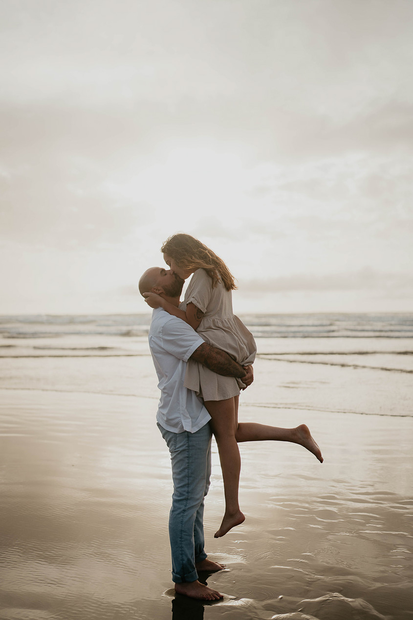 a newly engaged couple kissing while standing barefoot in Cannon beach. 