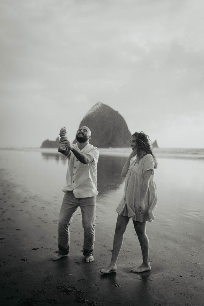 a newly engaged couple popping open a bottle of champagne at Cannon Beach with Haystack Rock in the background.  