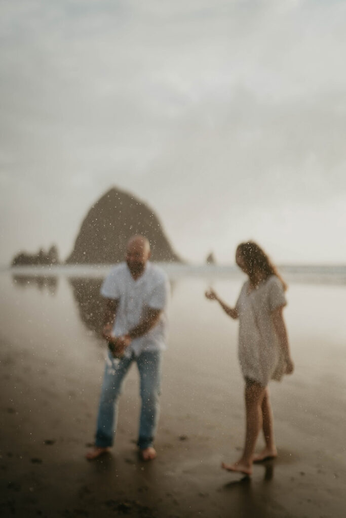 a newly engaged couple popping open a bottle of champagne with Haystack Rock in the background.  