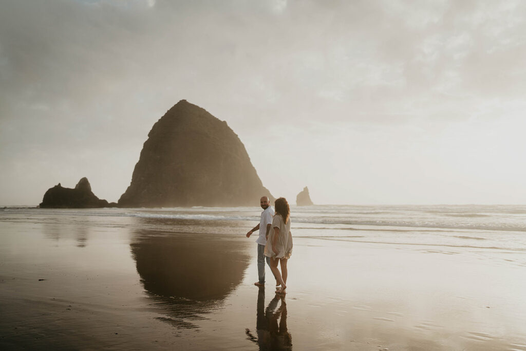 a newly engaged couple walking with Haystack Rock in the background. 