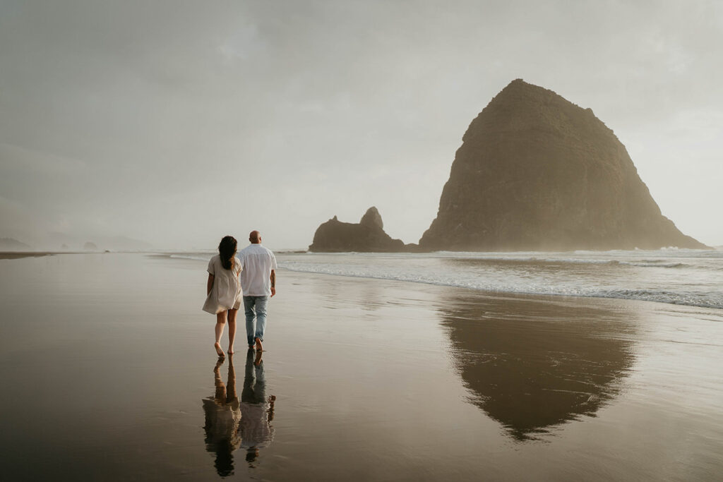a newly engaged couple walking along Cannon Beach with Haystack Rock in the background. 