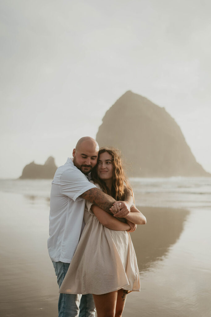 A newly engaged couple hugging at Cannon Beach with Haystack Rock in the background. 