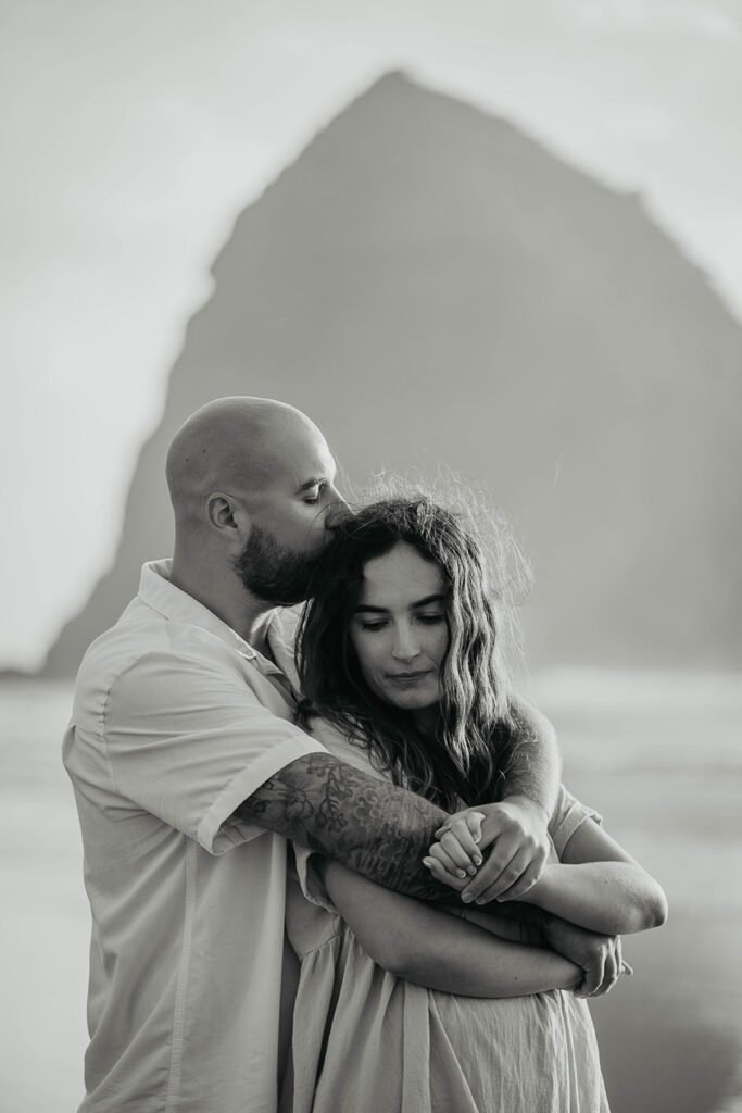 A newly engaged couple hugging and kissing at Cannon Beach with Haystack Rock in the background. 