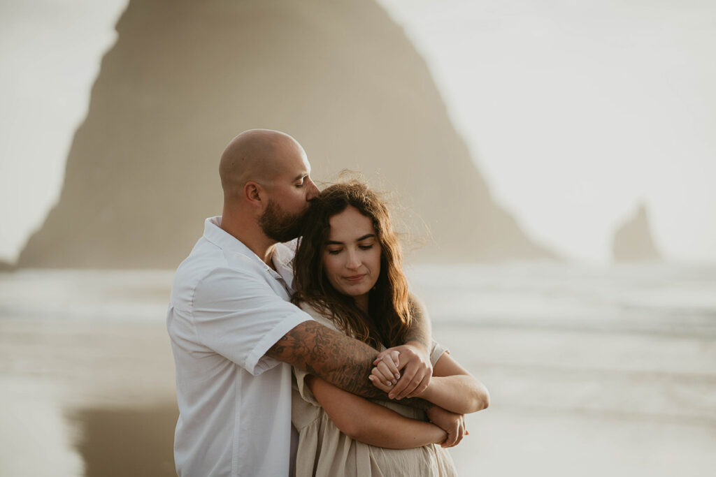 A newly engaged couple hugging and kissing at Cannon Beach with Haystack Rock in the background. 