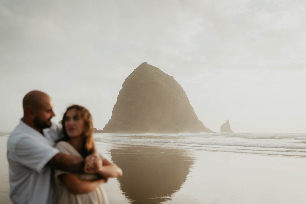 A newly engaged couple staring into each other's eyes at Cannon Beach with Haystack Rock in the background. 