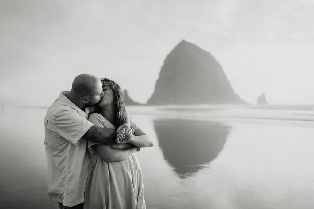 A newly engaged couple kissing at Cannon Beach with Haystack Rock in the background. 