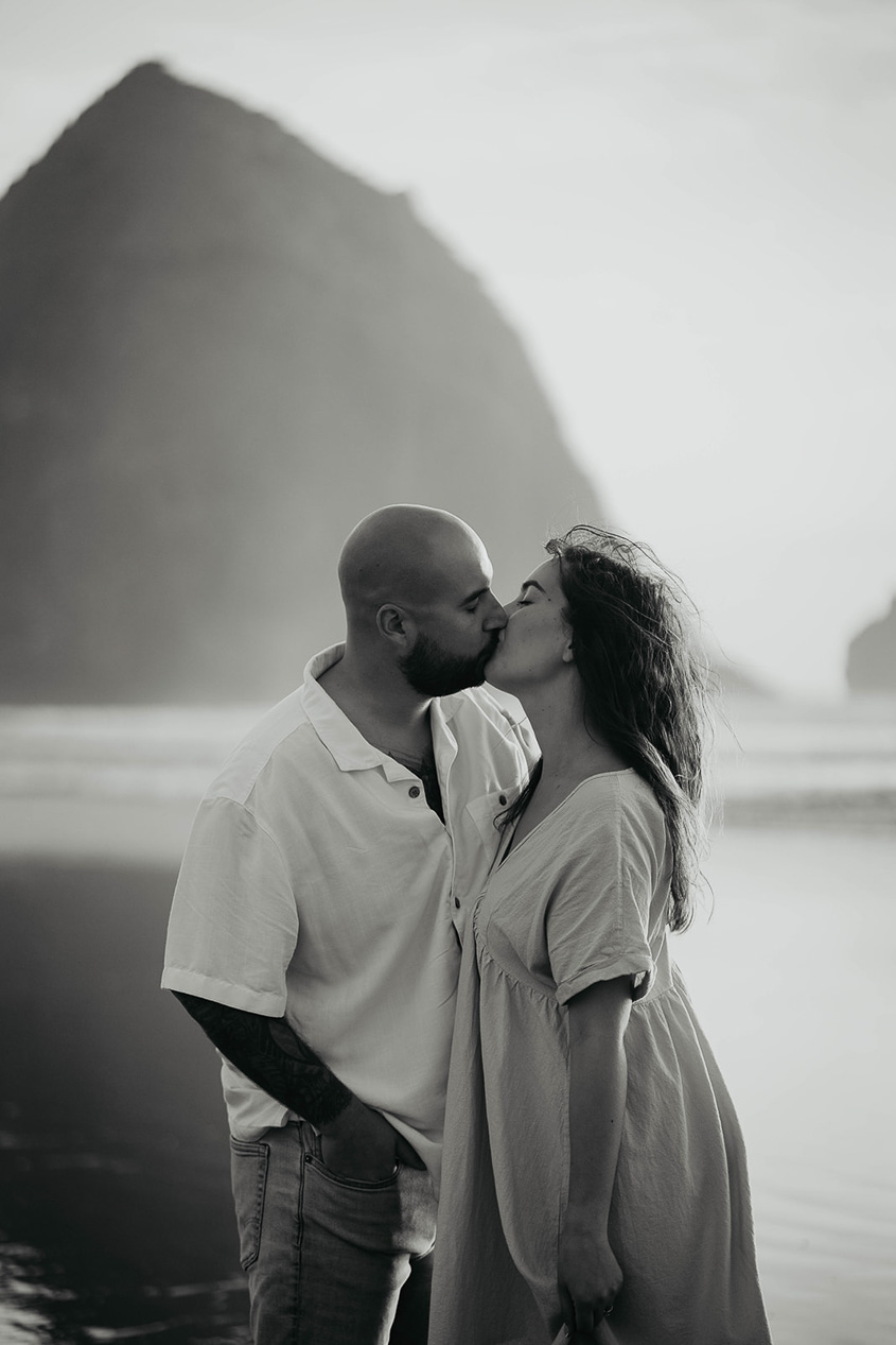a newly engaged couple kissing at Cannon Beach with Haystack Rock in the background. 