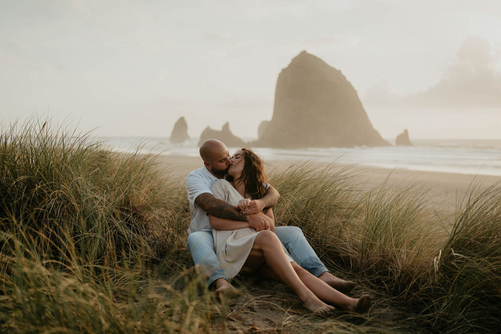 a couple sitting in beach grass kissing with Haystack Rock and Cannon Beach behind them. 