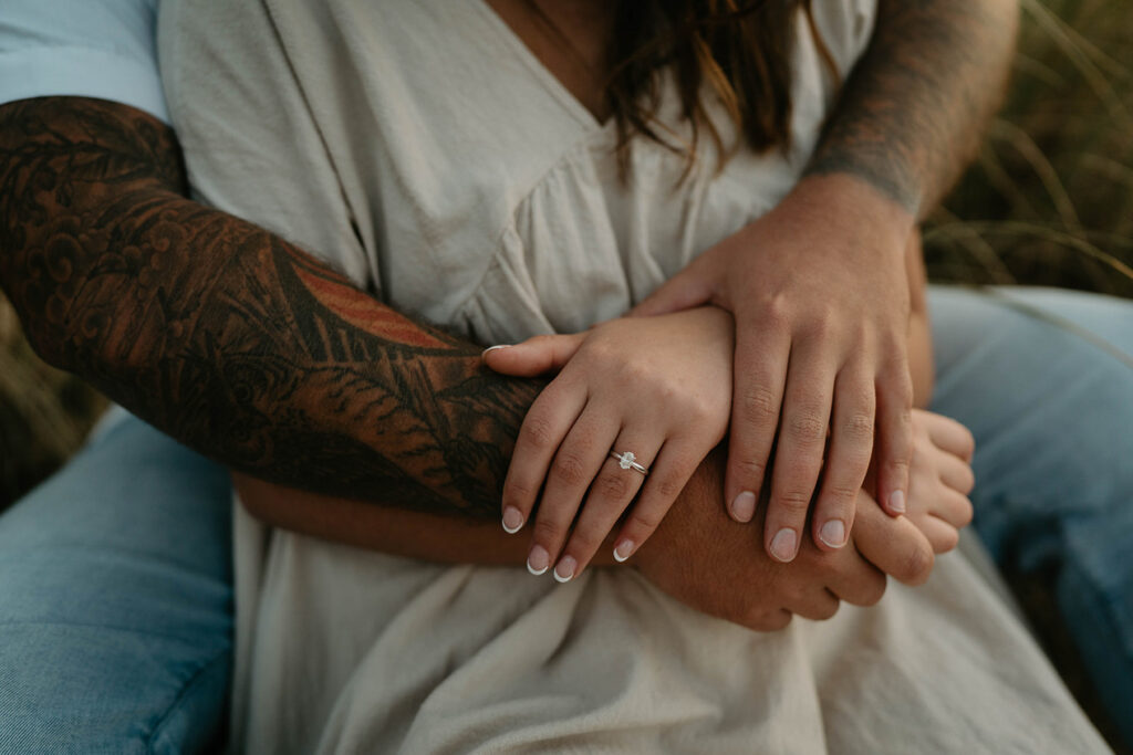 a couple sitting in beach grass, with the fiancee showing off her new engagement ring. 