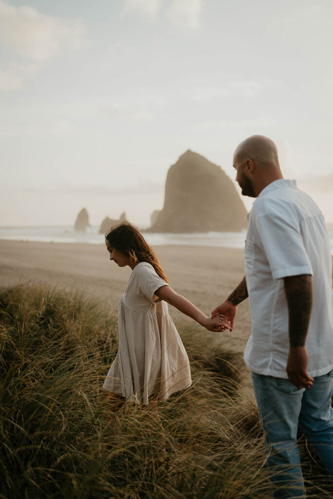a newly engaged couple walking among beach grass with Haystack Rock in the background. 