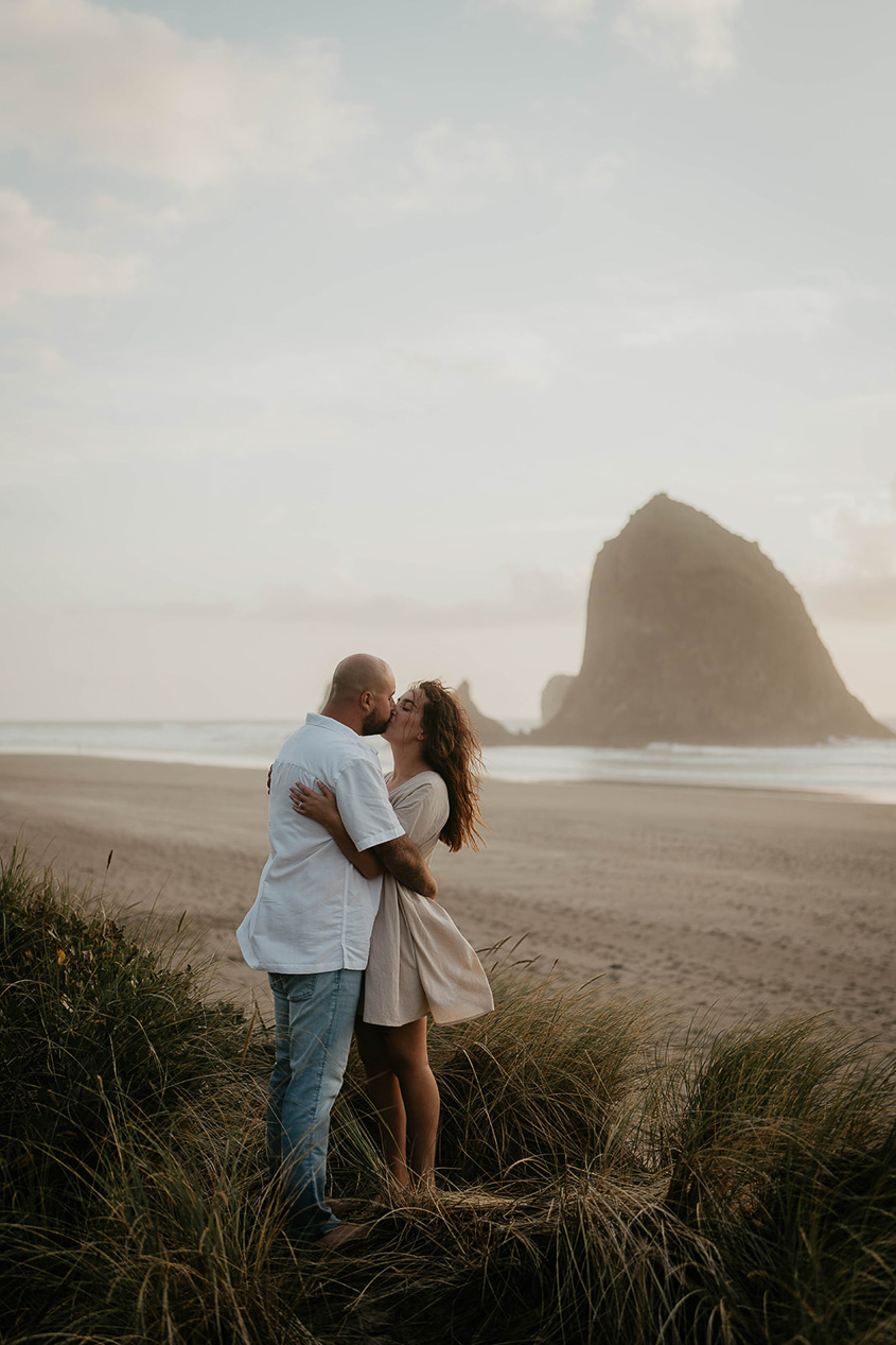 a newly engaged couple kissing among beach grass with Haystack Rock in the background. 