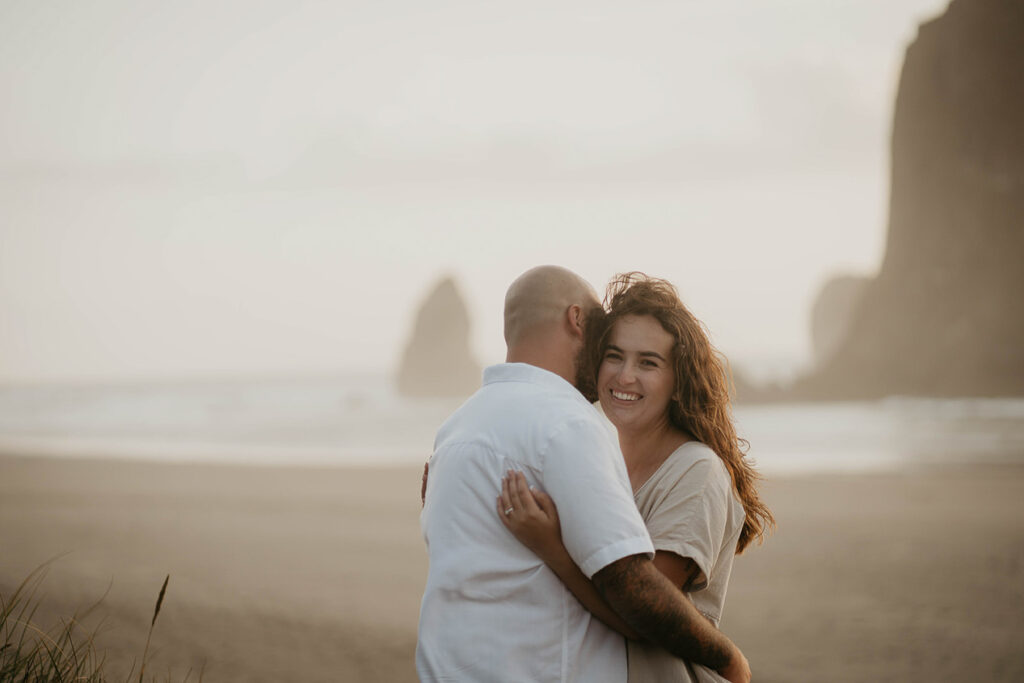 a newly engaged couple holding each other, one of them smiling brightly, with Haystack Rock in the background. 