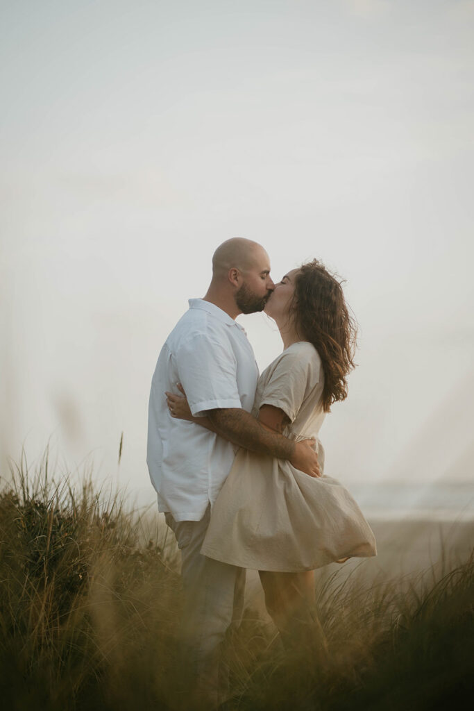 a newly engaged couple kissing among beach grass with the Pacific Ocean in the background. 