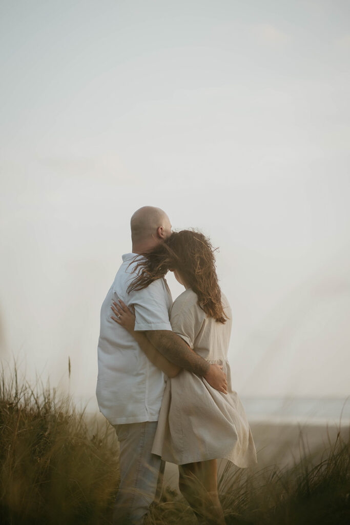 a newly engaged couple holding each other among beach grass and looking out to the Pacific Ocean. 