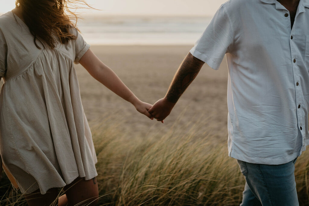 a newly engaged couple holding hands while walking among beach grass with the Pacific Ocean in the background. 