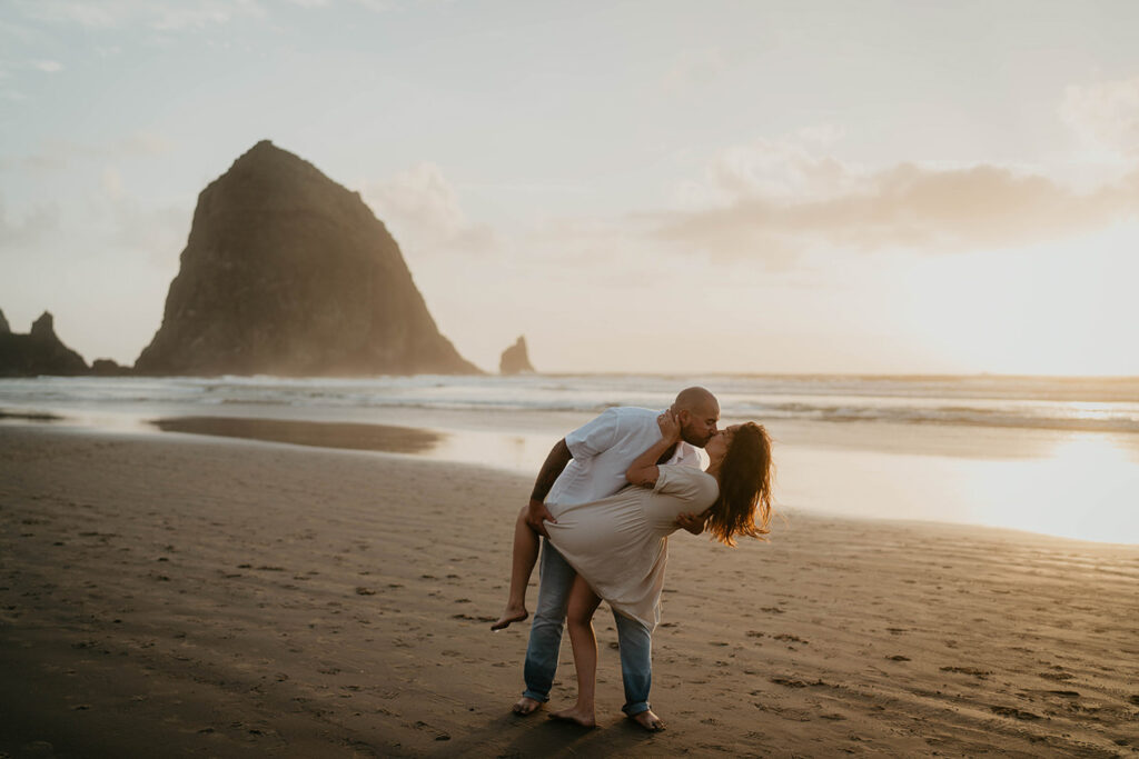 a newly engaged couple kissing as the sun is setting while standing on Cannon Beach with  Haystack rock in the background. 