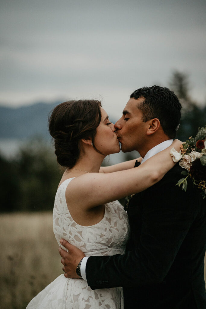 the newlyweds kissing with a field and pine trees in the background. 
