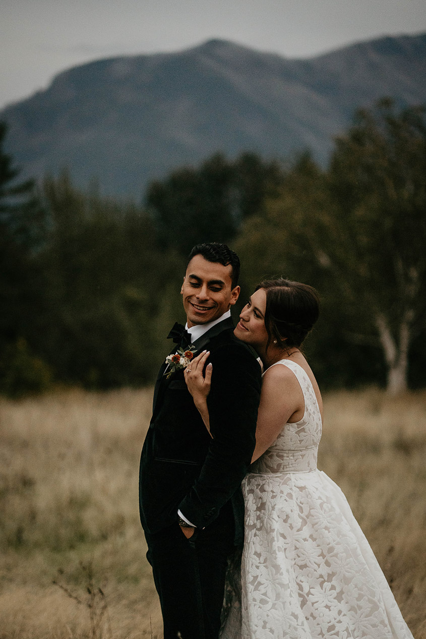 the newlyweds hugging with a field and pine trees in the background. 