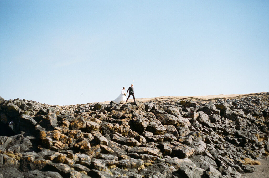 Bride and groom hold hands while walking across the rocks for their film wedding photos