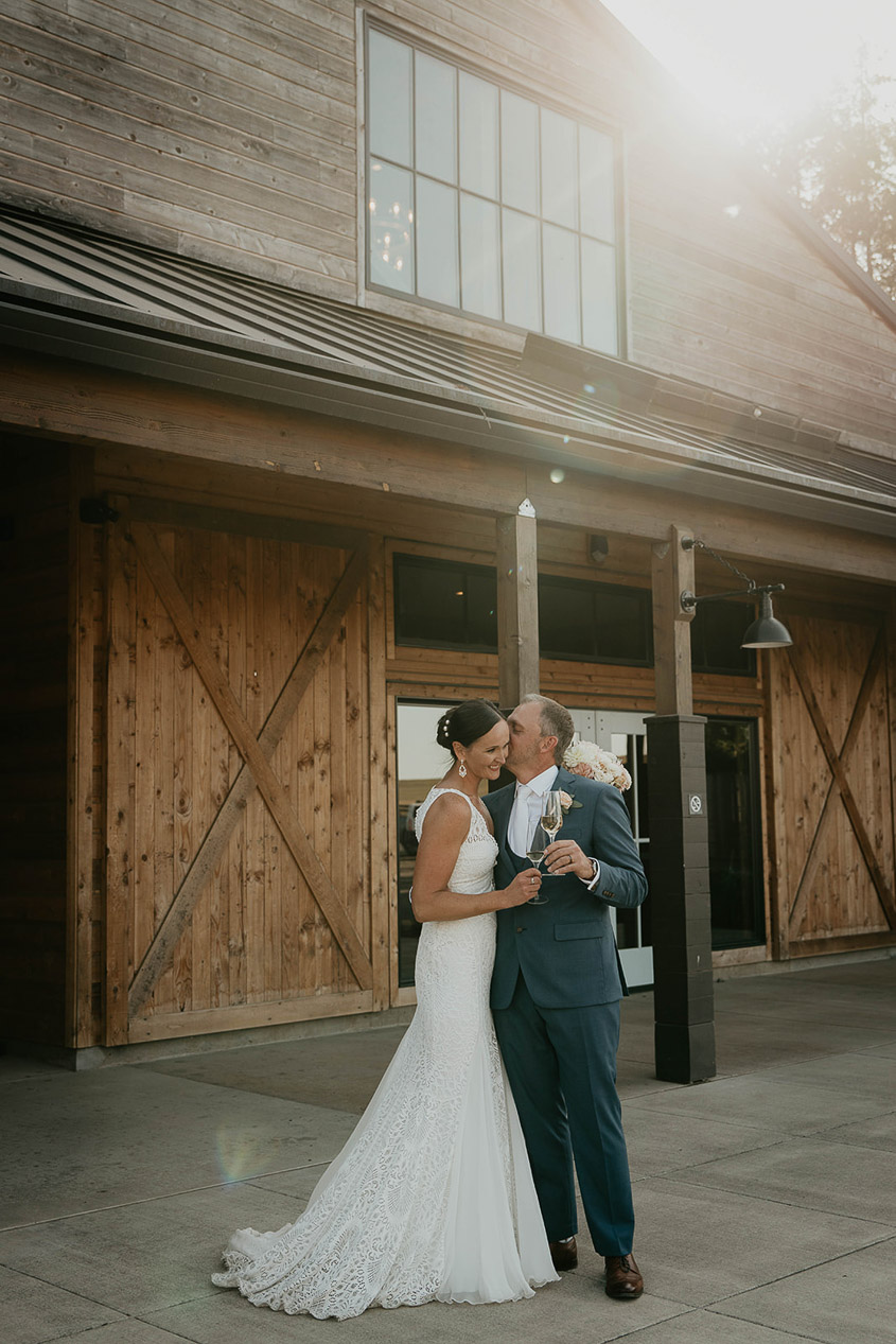 The bride and groom kissing and holding champagne by a wooden barn. 