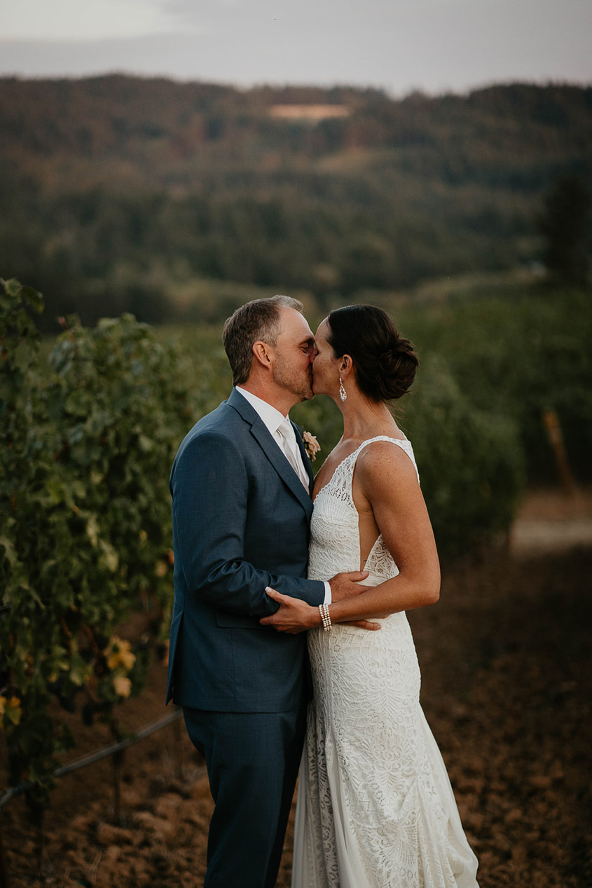 the bride and groom kissing by a vineyard at sunset. 