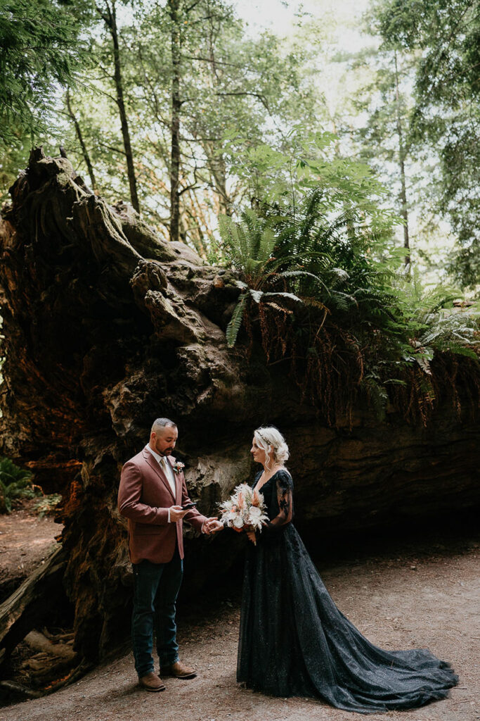 a couple holding flowers in front of a fallen tree trunk for their Redwoods elopement. 
