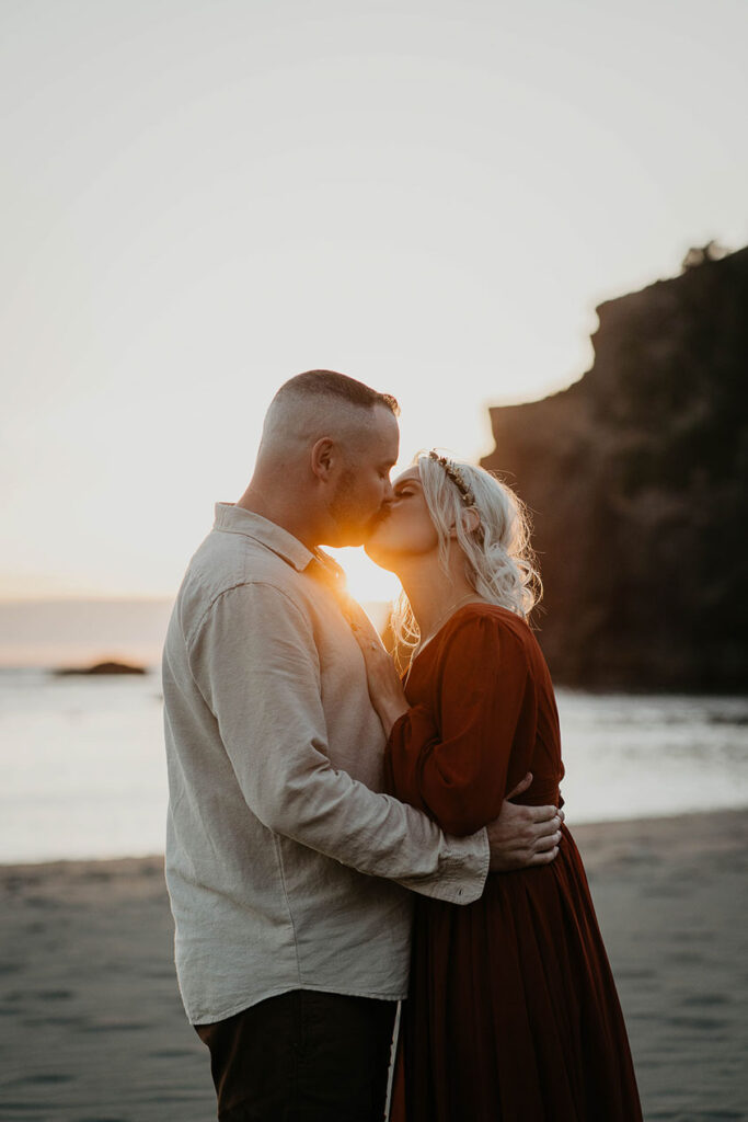 The husband and wife kissing as the sun sets over the Pacific Ocean. 