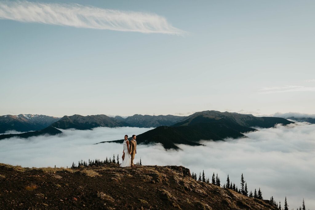 Bride and groom stand back to back on a mountaintop in Olympic National Park surrounded by clouds