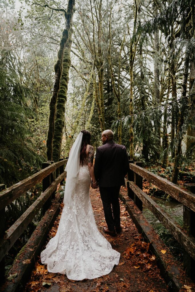 Bride and groom hold hands while walking across a wood bridge at their Washington elopement in the Hoh Rainforest