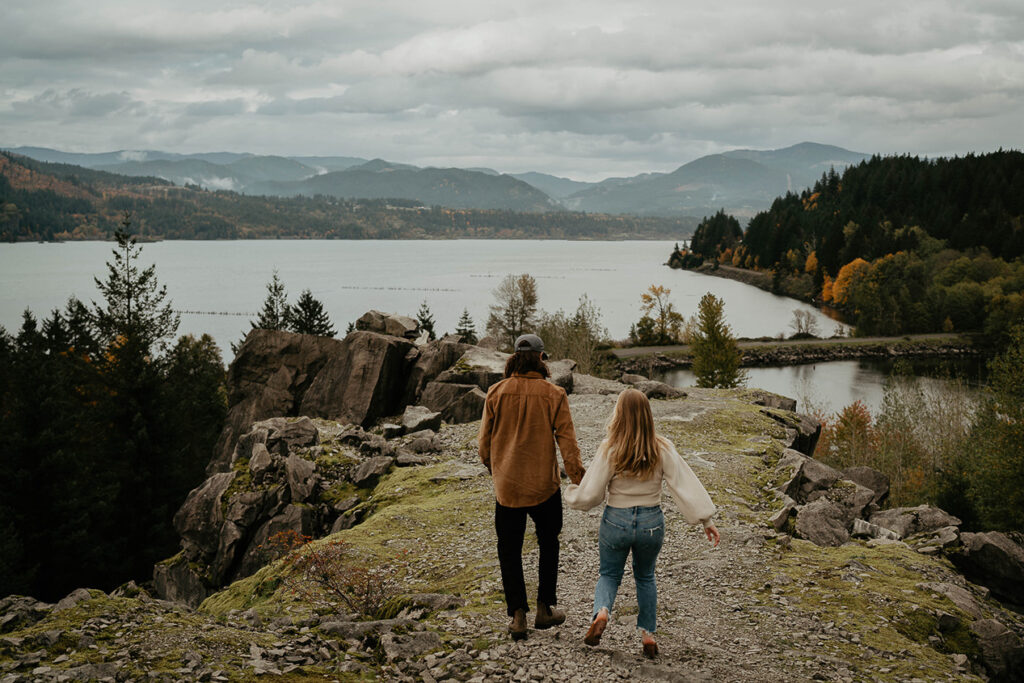 a newly engaged couple holding hands and walking with the Columbia River Gorge in the background. 